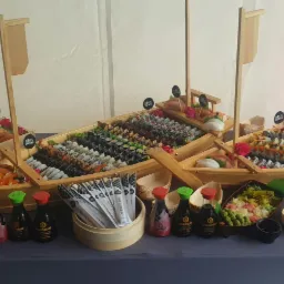 Bogato zaopatrzony stół z sushi ułożonym w dekoracyjnych łodziach, uzupełniony o sos sojowy, pałeczki i małe miseczki, całość sygnowana logo 'KOKU Sushi'.