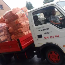 Biała ciężarówka K2500 z czerwonymi akcentami, wypełniona workami z pociętym drewnem, kierowca w kabinie, numer telefonu na drzwiach, tło miejskie.