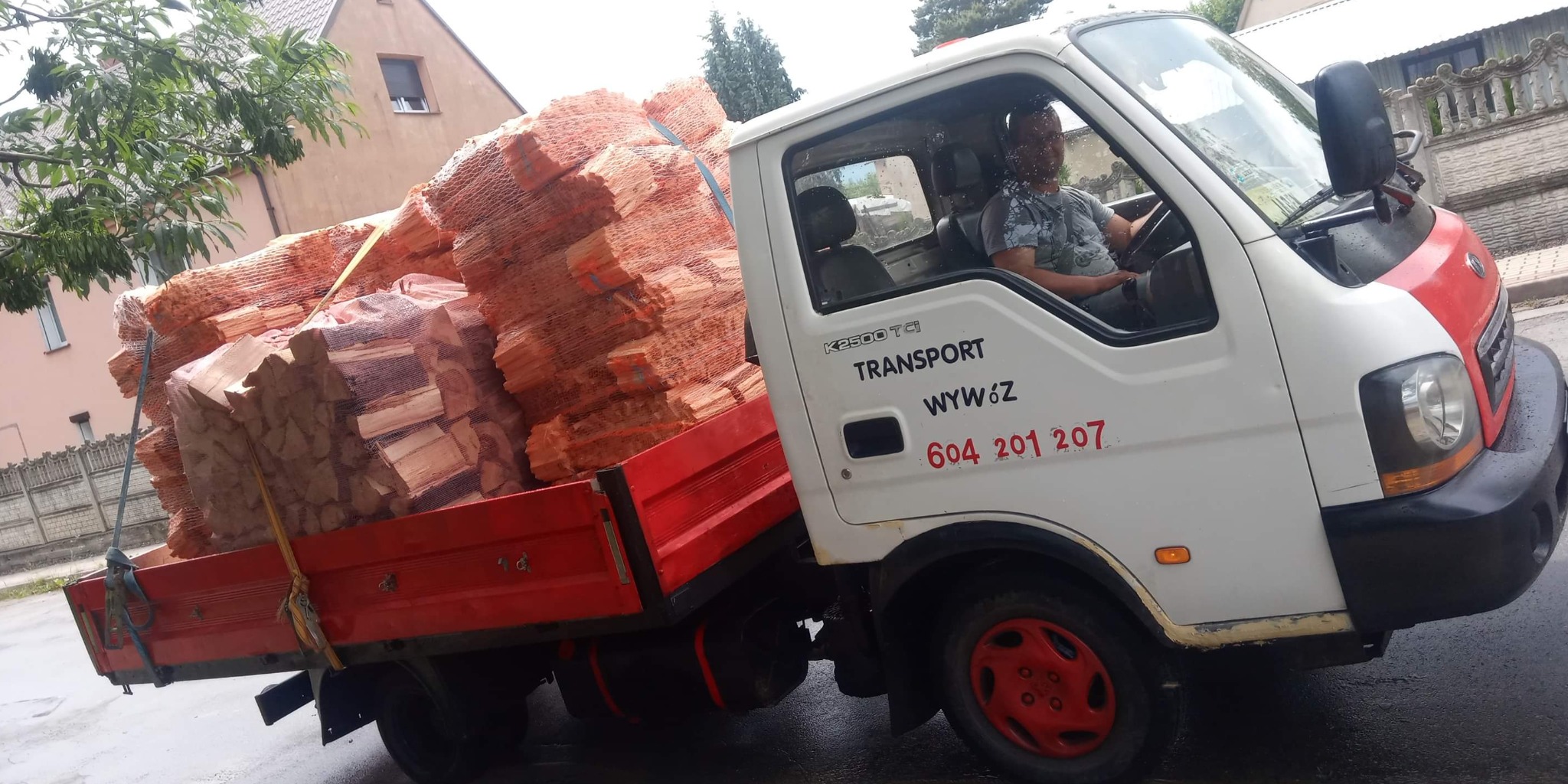 Biała ciężarówka K2500 z czerwonymi akcentami, wypełniona workami z pociętym drewnem, kierowca w kabinie, numer telefonu na drzwiach, tło miejskie.