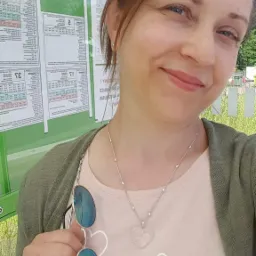 Portret uśmiechniętej kobiety w jasnej bluzce z napisem i naszyjnikiem z sercem, stojącej przy zielonym przystanku autobusowym z rozkładami jazdy.