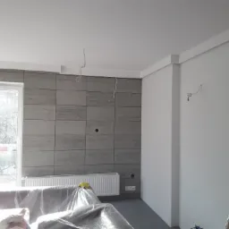 Ściana z szarych płytek imitujących kamień, biały grzejnik, zakryte folią meble, widoczne przewody elektryczne wystające ze ściany i sufitu, fragment drabiny malarskiej w rogu, okno z widokiem...