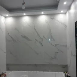 Wykończona ściana łazienki z białych płytek imitujących marmur, z widocznym grzejnikiem drabinkowym i wystającą żarówką na kablu.