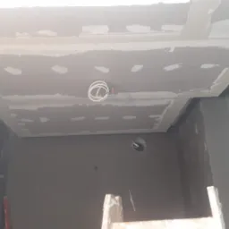 Niedokończone prace przygotowawcze do malowania: sufit z płyt gipsowo-kartonowych z widocznymi łączeniami i kablami oświetleniowymi, ściany w szarym kolorze, fragment drabiny w dolnej części kadru.