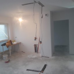 Mateusz Klass wykończenia wnętrz - Pokój w trakcie prac wykończeniowych: widoczne przewody elektryczne zwisające z sufitu, przygotowane ściany, prowizoryczne biurko z ubraniami i narzędzia na podłodze.