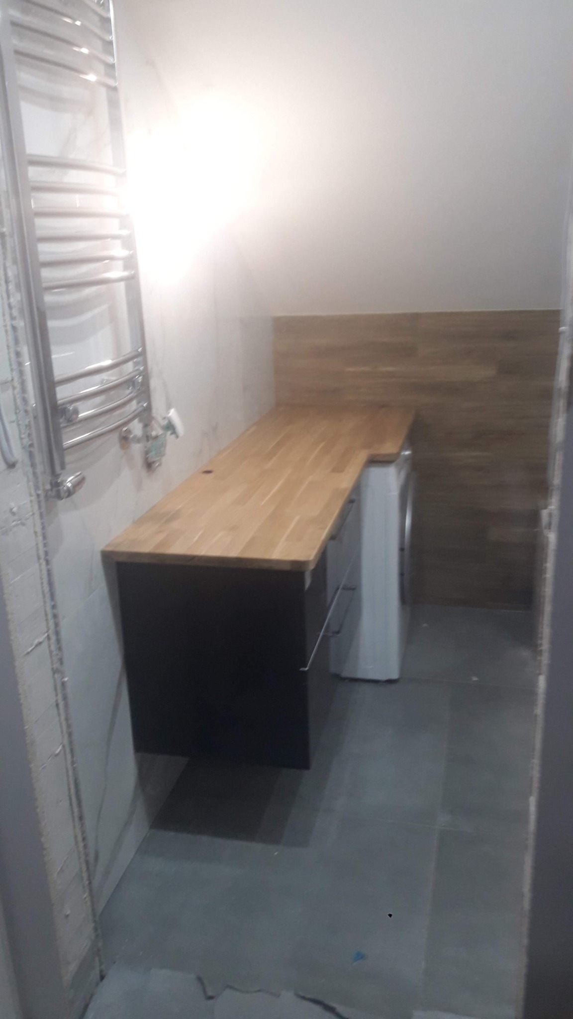 Blat z jasnego drewna nad pralką i szafką z szufladami w łazience z szarą podłogą i grzejnikiem drabinkowym na ścianie wyłożonej płytkami.