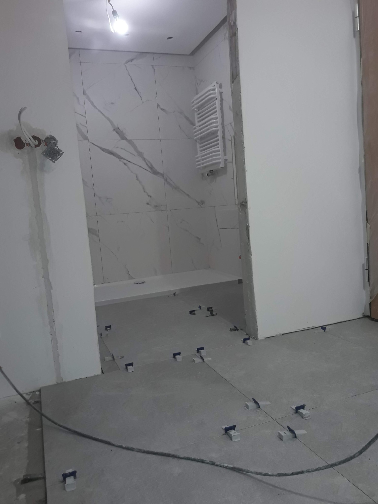 Wnętrze łazienki w trakcie prac w Tokarach: ułożone szare płytki podłogowe z systemem poziomowania, białe płytki imitujące marmur na ścianach, widoczny brodzik i grzejnik drabinkowy.