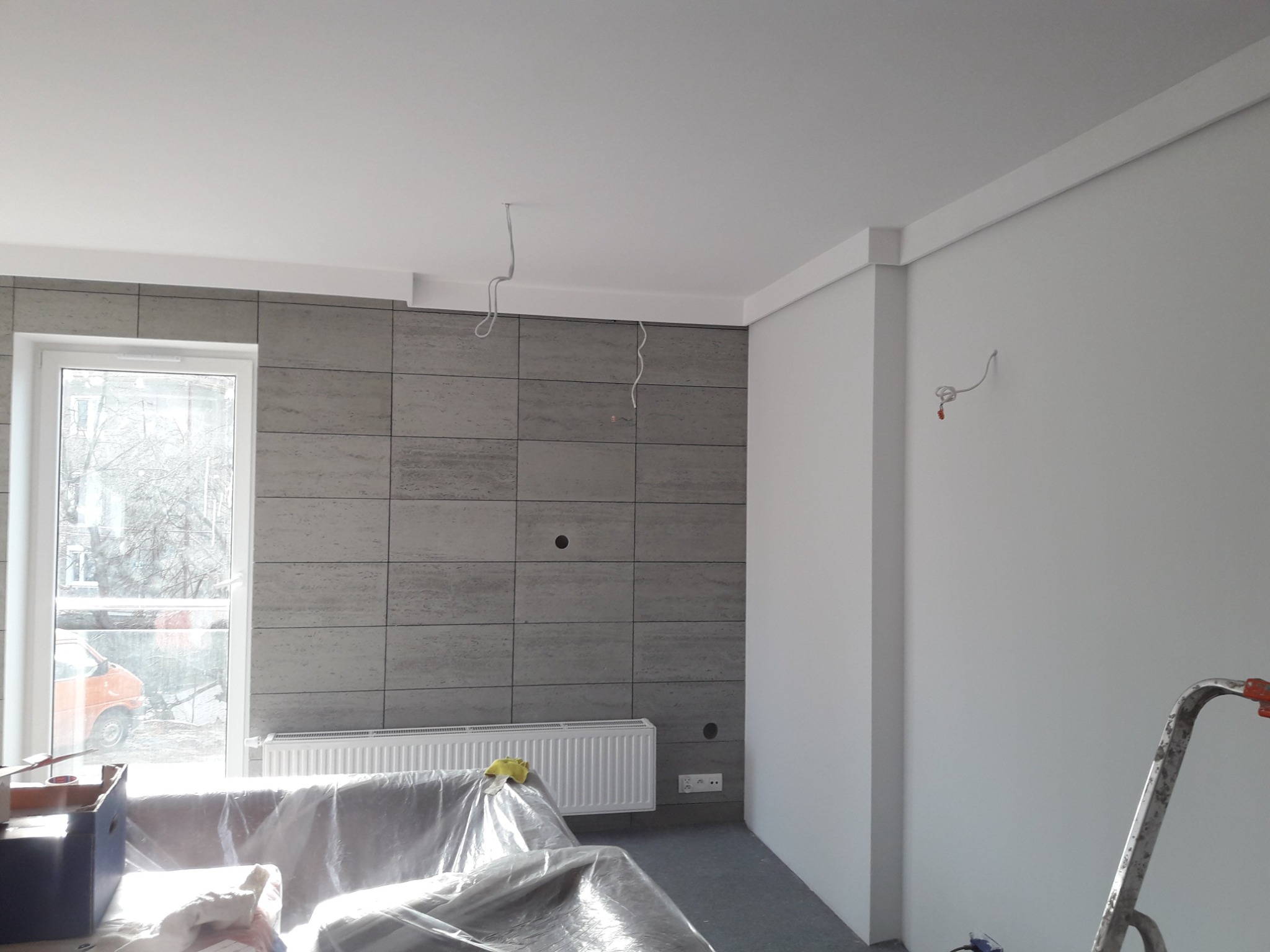 Ściana z szarych płytek imitujących kamień, biały grzejnik, zakryte folią meble, widoczne przewody elektryczne wystające ze ściany i sufitu, fragment drabiny malarskiej w rogu, okno z widokiem...