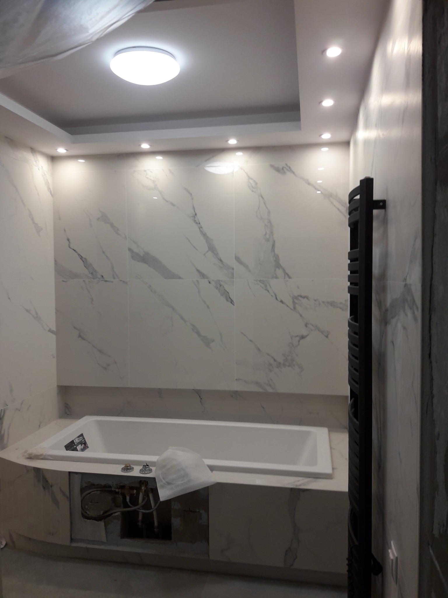 Wanna w obudowie z marmurowymi płytkami, widoczne przyłącza hydrauliczne w otworze rewizyjnym, grzejnik drabinkowy na ścianie z gładzią.