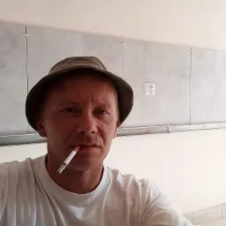 Glazurnik w kapeluszu z papierosem w ustach na tle ściany z szarymi płytkami w trakcie montażu, widoczne krzyżyki dystansowe i puszki elektryczne.