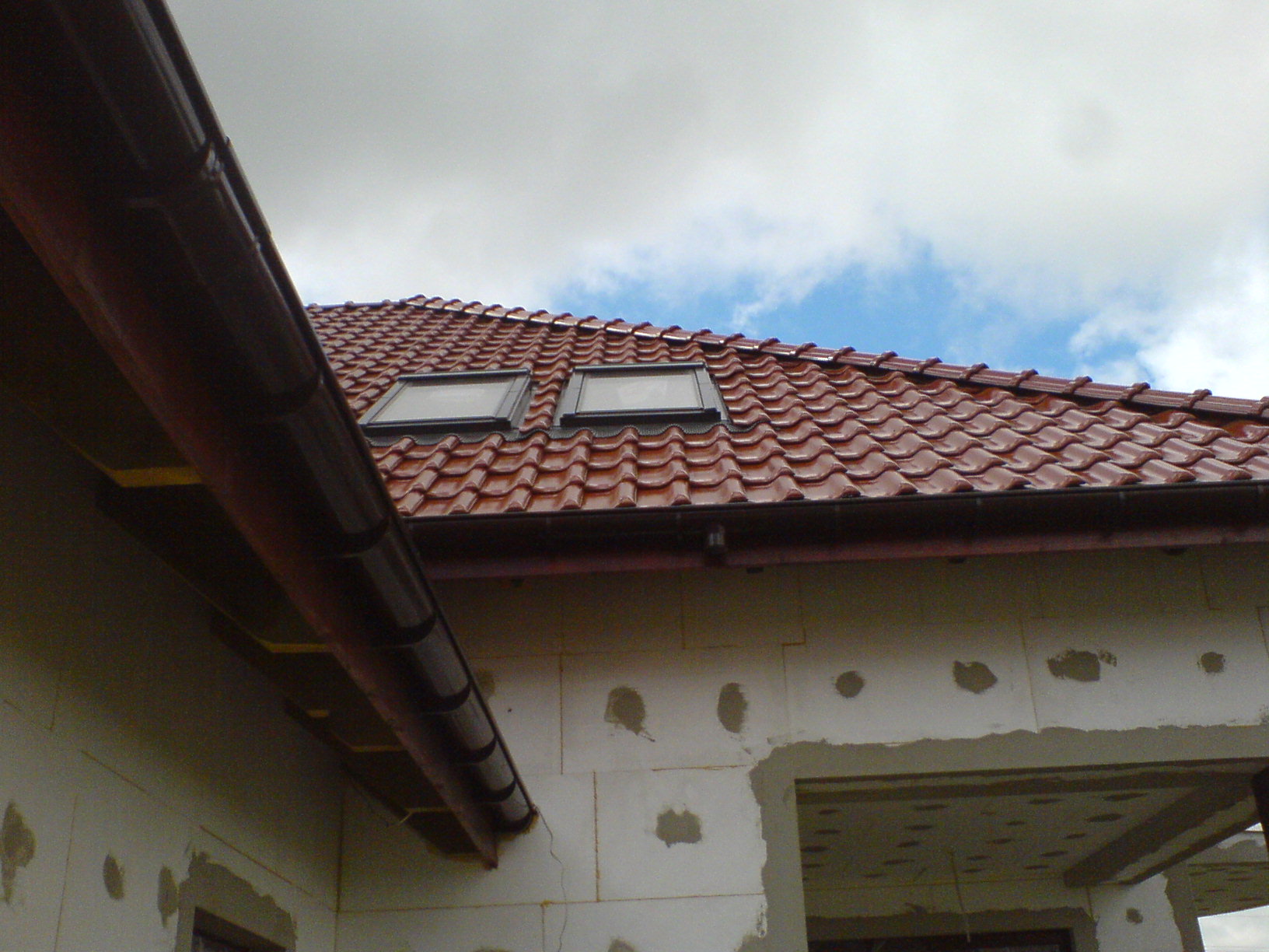 Czerwona dachówka ceramiczna z dwoma oknami dachowymi, brązowa rynna, ściana budynku w trakcie ocieplania, błękitne niebo z chmurami w tle.