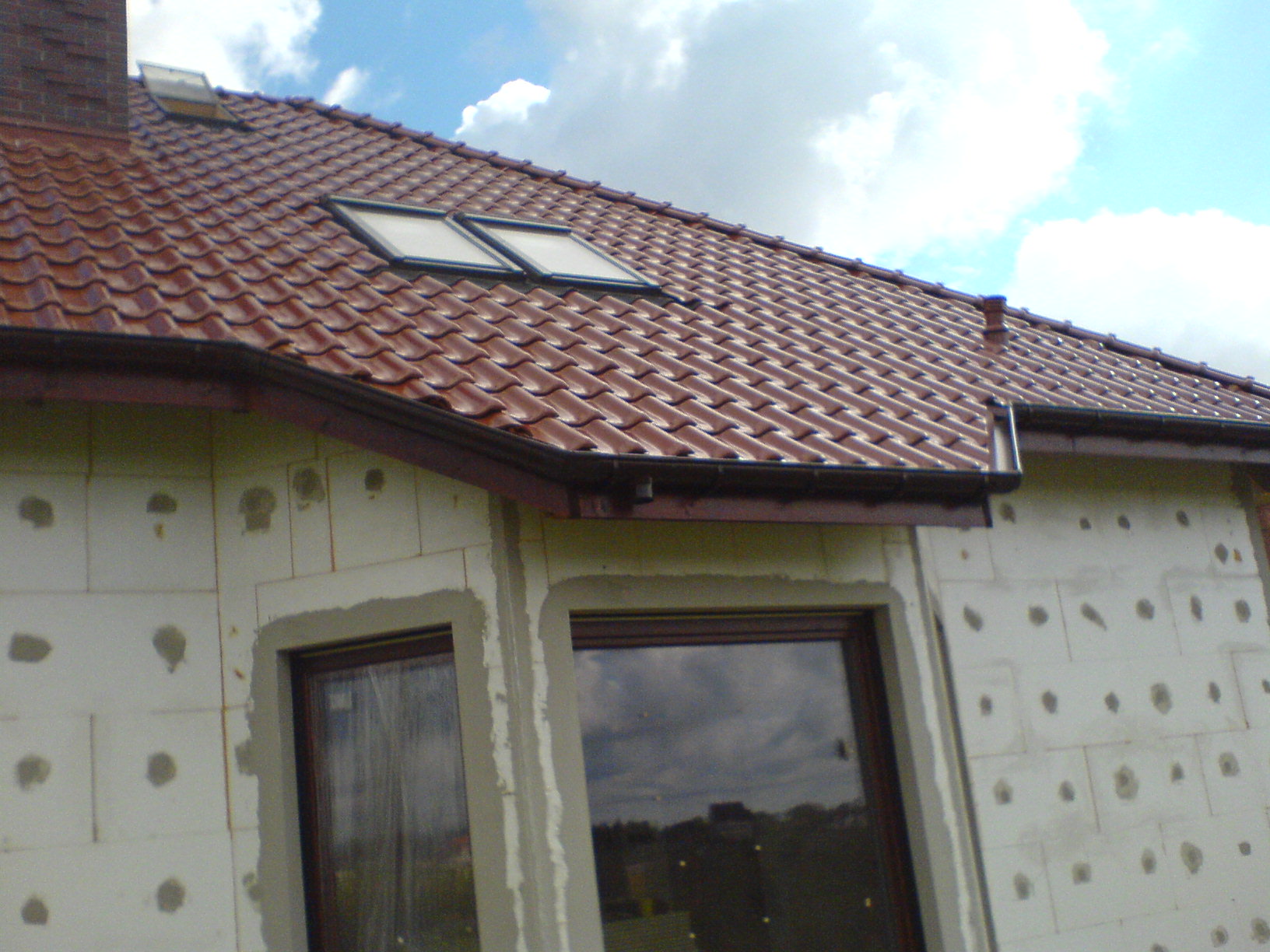 Nowy dach z czerwoną dachówką ceramiczną i oknami połaciowymi na budynku w trakcie ocieplania styropianem.