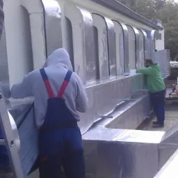 Montaż systemu wentylacyjnego na dachu budynku, widoczni dwaj pracownicy w strojach roboczych, jeden z nich stoi na drabinie trzymając element instalacji, w tle budynki i drzewa.