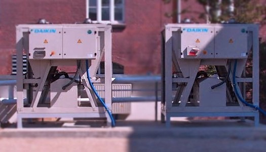 Dwa agregaty chłodnicze Daikin na dachu budynku z cegły, połączone rurami, widoczne elementy instalacji i okno w tle.