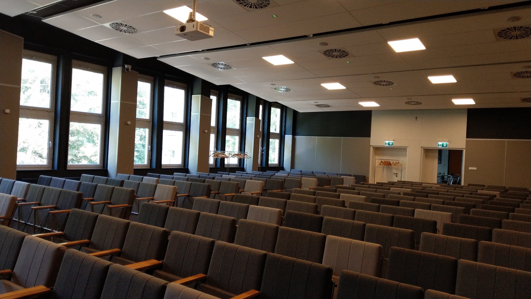 Sala konferencyjna z rzędami szarych krzeseł, widoczne okna z naturalnym światłem, projektor podwieszony do sufitu oraz kratki wentylacyjne. Na ścianie widoczne drzwi z oznaczeniami ewakuacyjnymi.