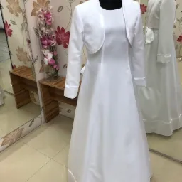 Biała sukienka komunijna z bolerkiem na manekinie, widoczny detal wykończenia satynową lamówką, w tle druga sukienka i dekoracyjna ściana.