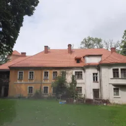 Odnowiony dach z czerwonej dachówki na zabytkowym, częściowo odrestaurowanym budynku z żółtą i białą elewacją, widoczne kominy i fragmenty zieleni.