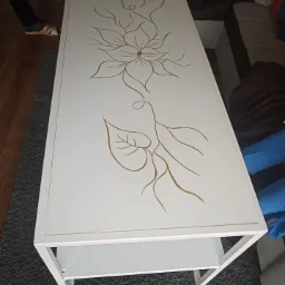 Biały stolik kawowy z metalową ramą i blatem zdobionym złotym, florystycznym motywem, widziany z góry.