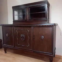 Ciemnobrązowy, drewniany kredens z nadstawką przeszkloną, zdobiony rzeźbieniami, ustawiony w pokoju na drewnianej podłodze, obok pufa w podobnej tonacji.