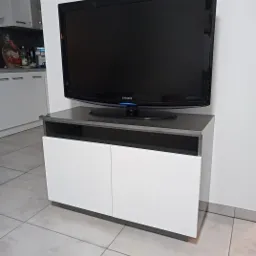 Nowoczesna szafka RTV z białymi frontami i szarym blatem, na której stoi telewizor Samsung, ustawiona na jasnej płytce podłogowej.