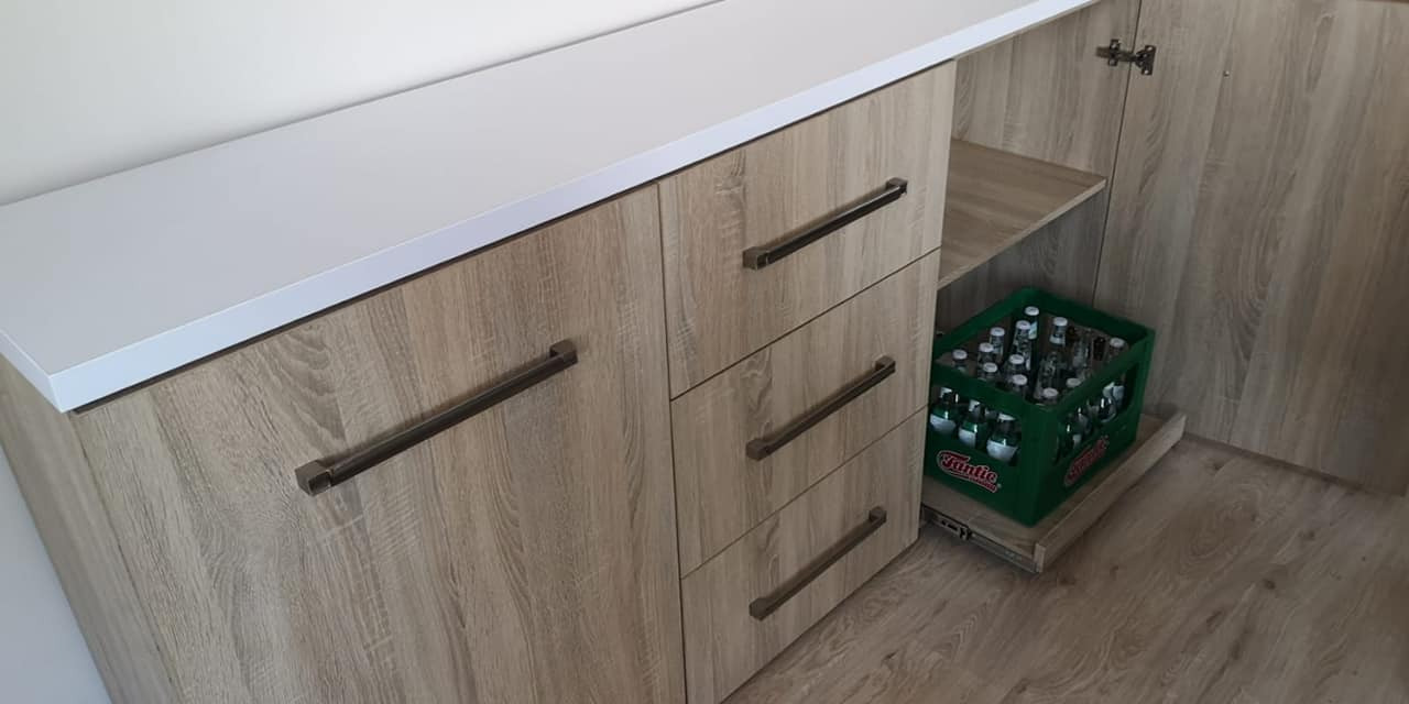 Jasna komoda z trzema szufladami i szafką z blatem w kolorze białym, widoczna skrzynka z napojami na wysuwanej półce wewnątrz szafki.