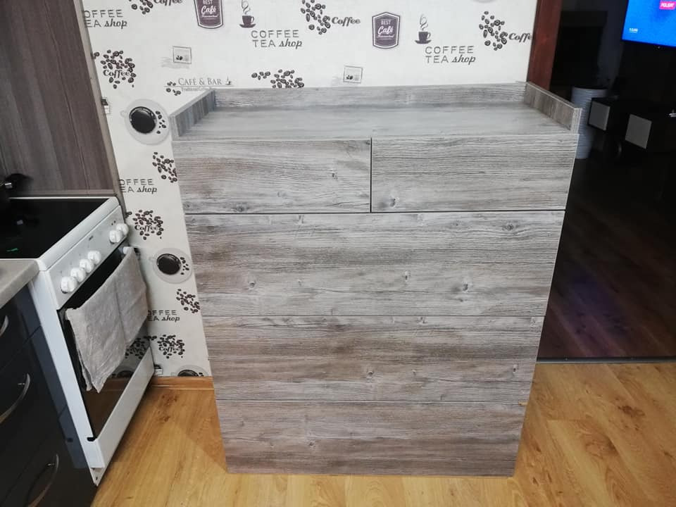 Niska komoda z szufladami w kuchni, wykończona okleiną imitującą jasne drewno, stojąca obok białej kuchenki, na tle ściany z tapetą w motywy kawowe.