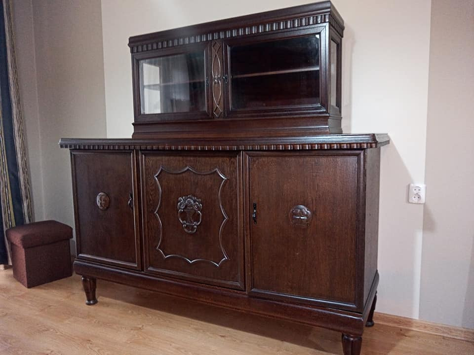 Ciemnobrązowy, drewniany kredens z nadstawką przeszkloną, zdobiony rzeźbieniami, ustawiony w pokoju na drewnianej podłodze, obok pufa w podobnej tonacji.