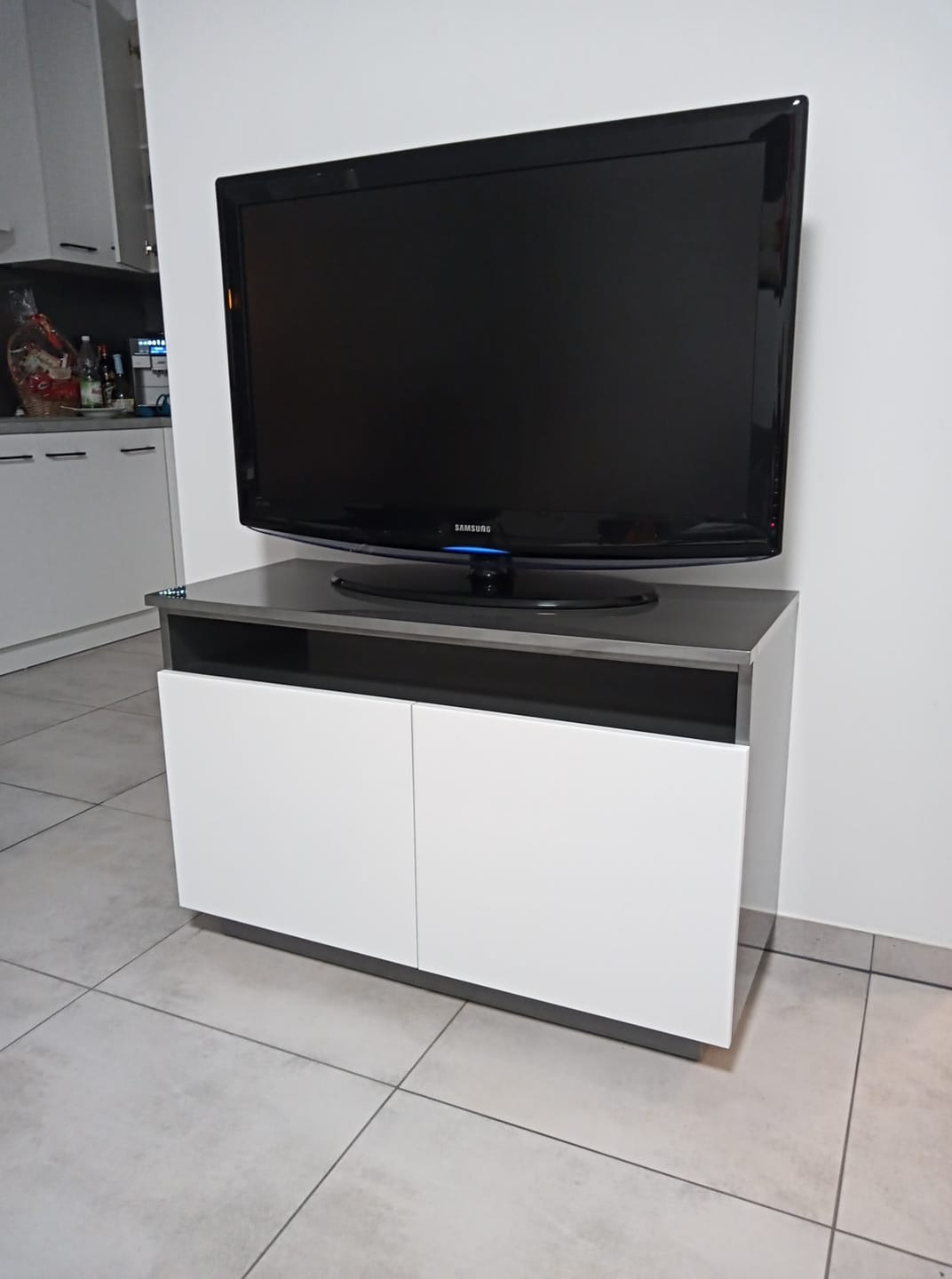 Nowoczesna szafka RTV z białymi frontami i szarym blatem, na której stoi telewizor Samsung, ustawiona na jasnej płytce podłogowej.