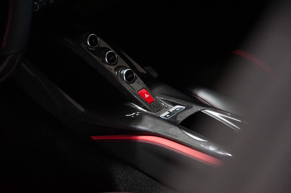 Szczegółowe ujęcie konsoli środkowej samochodu sportowego z włókna węglowego z czerwonymi akcentami, prezentujące przyciski sterujące i charakterystyczny czerwony przycisk awaryjny.