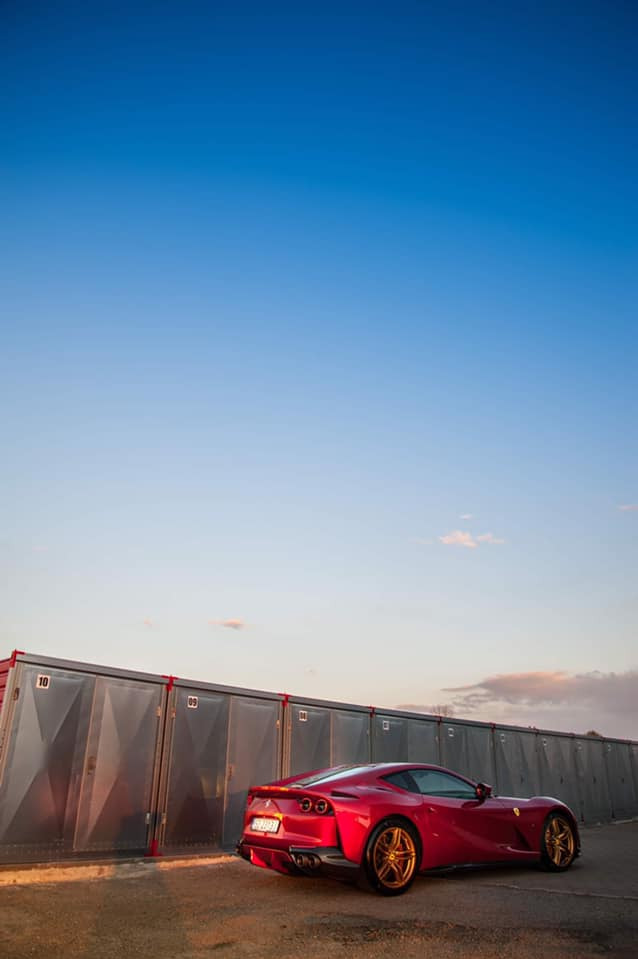 Czerwone Ferrari 812 Superfast z złotymi felgami zaparkowane przed rzędem szarych garaży kontenerowych w promieniach zachodzącego słońca.