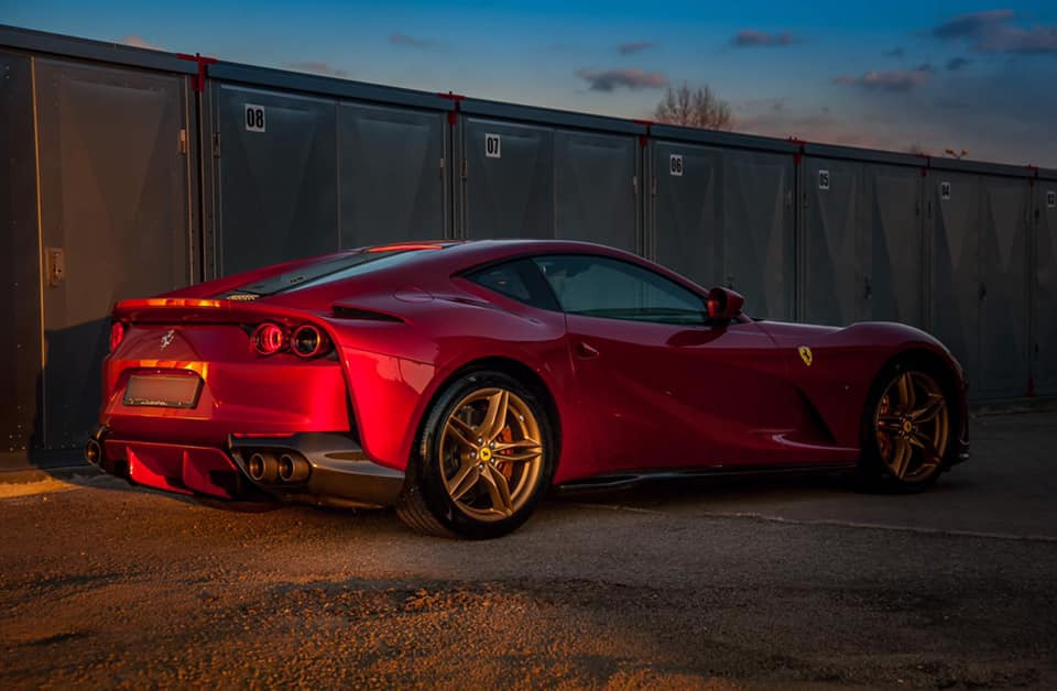 Czerwone Ferrari 812 Superfast zaparkowane przed szarymi garażami, widok z tyłu, złote felgi, zachodzące słońce.