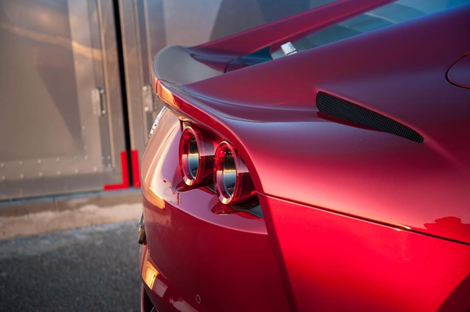 Czerwony sportowy samochód z widocznymi okrągłymi tylnymi światłami i spojlerem, zaparkowany przed metalową bramą garażową.