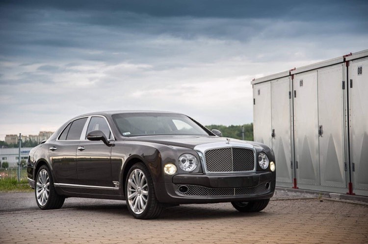 Ciemnoszary luksusowy sedan Bentley Mulsanne zaparkowany na brukowanym placu przed rzędem srebrnych kontenerów, pochmurne niebo w tle.