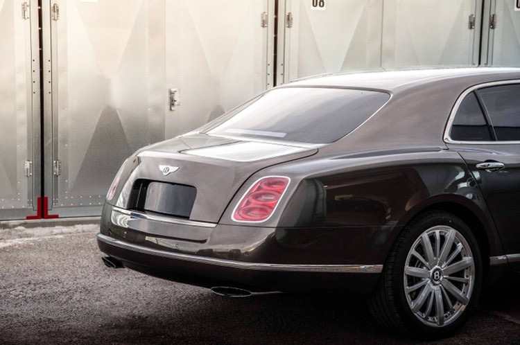 Brązowy luksusowy samochód Bentley Mulsanne z charakterystycznymi tylnymi światłami i chromowanymi detalami, zaparkowany przed szarymi metalowymi drzwiami garażowymi z numerem '08' u góry.