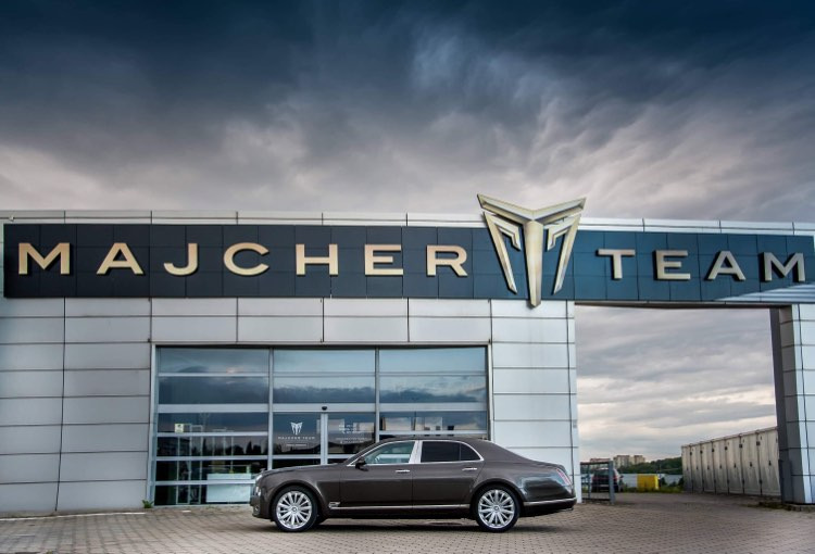 Brązowy luksusowy samochód Bentley Mulsanne zaparkowany przed budynkiem z napisem 'MAJCHER TEAM' i dużym srebrnym logo na tle pochmurnego nieba.