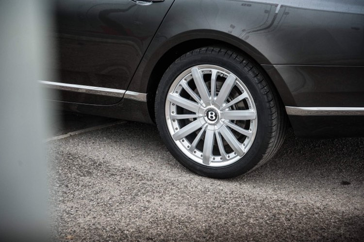 Fragment czarnego samochodu marki Bentley z widocznym srebrnym felgi z logo firmy na tle szarego asfaltu.