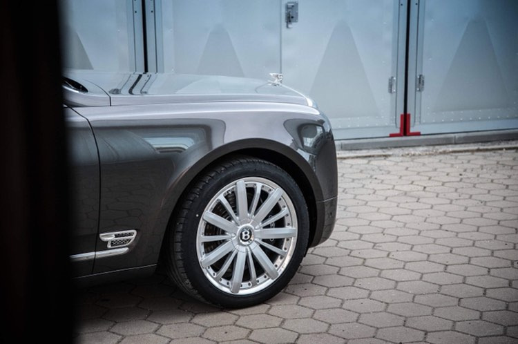 Szary luksusowy samochód Bentley z chromowanymi felgami i charakterystyczną figurką na masce, zaparkowany na kostce brukowej przed szarą ścianą z metalowymi drzwiami.