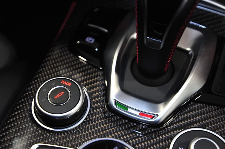 Szczegółowe ujęcie konsoli środkowej samochodu z włókna węglowego, z widocznym selektorem trybów jazdy z napisem 'RACE' i dźwignią zmiany biegów z czerwoną nicią oraz akcentami w kolorach włoskiej...