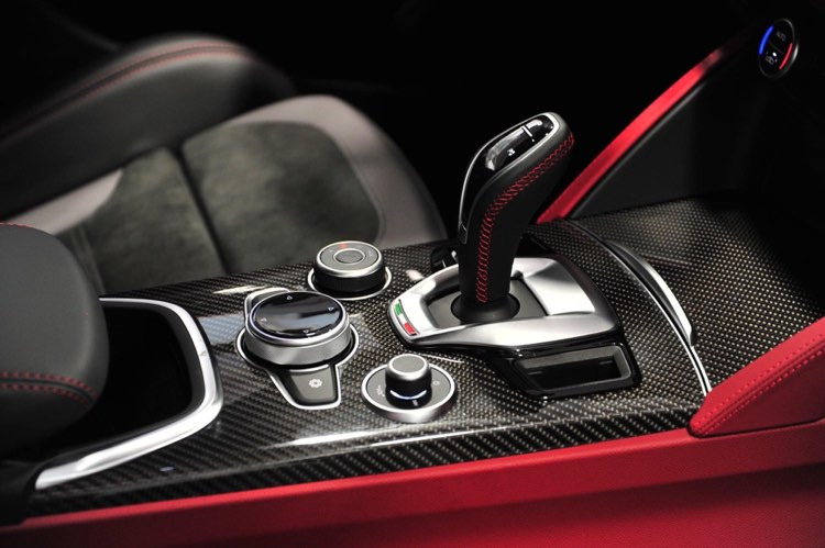 Detal wnętrza samochodu z włókna węglowego, dźwignia zmiany biegów z czerwoną nicią, srebrne pokrętła i czerwone akcenty tapicerki.