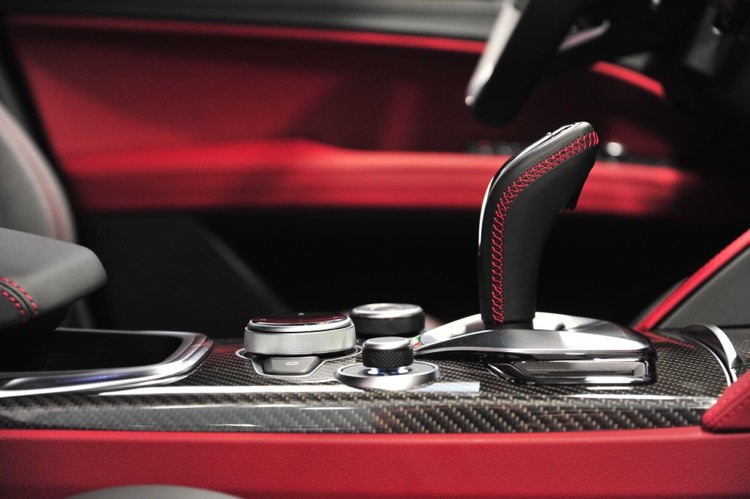 Wnętrze samochodu: dźwignia zmiany biegów z czerwoną nicią, pokrętła sterujące i deska rozdzielcza z włókna węglowego na tle czerwonej tapicerki.