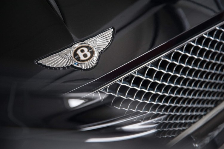 Błyszczące logo Bentley na czarnym lakierze maski samochodu, obok chromowanej kraty grilla.
