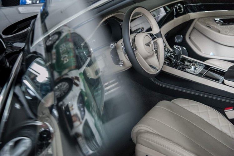 Wnętrze luksusowego samochodu z beżową skórzaną tapicerką, widoczne przez szybę z odbiciem otoczenia, kierownica z logo marki.