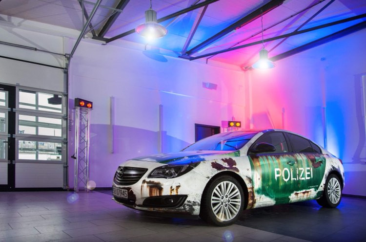 Samochód z napisem 'POLIZEI' w nietypowej, zardzewiałej stylistyce, oświetlony kolorowymi światłami w przestronnym garażu z podnoszoną bramą.