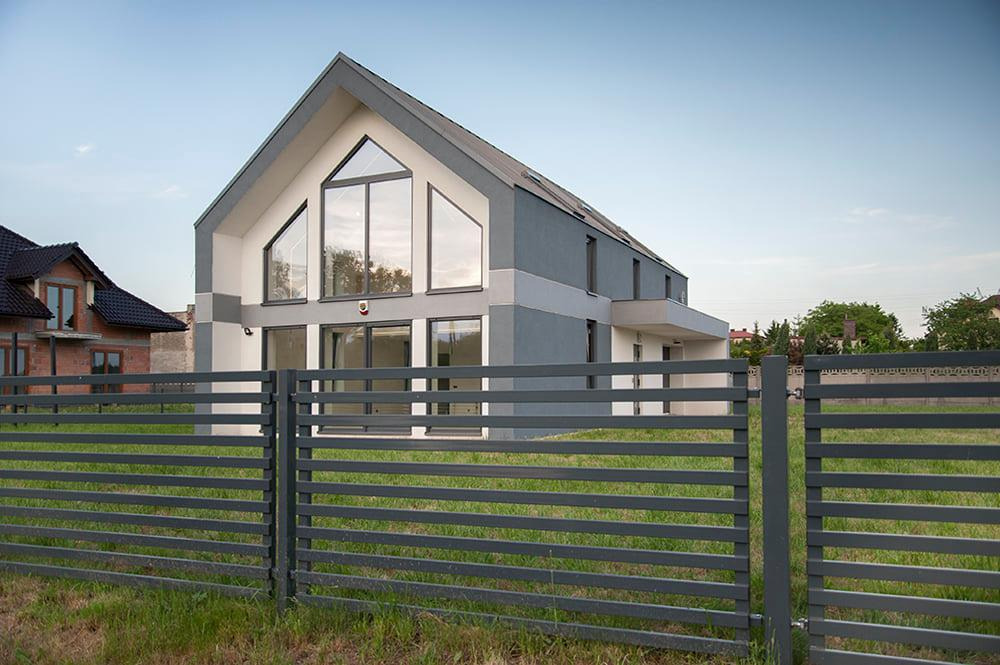 Nowoczesny dom jednorodzinny z dużymi oknami i szarą elewacją, widoczny zza metalowego ogrodzenia panelowego.