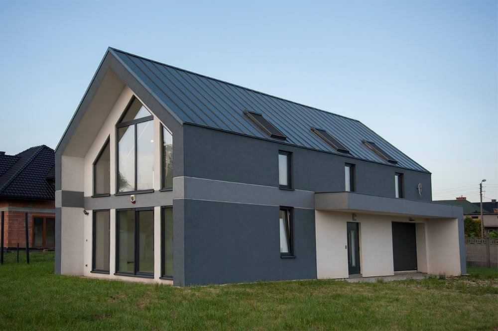 Nowoczesny dom jednorodzinny z grafitową elewacją i metalowym dachem, duże okna panoramiczne, widoczne okna dachowe, trawnik przed domem.
