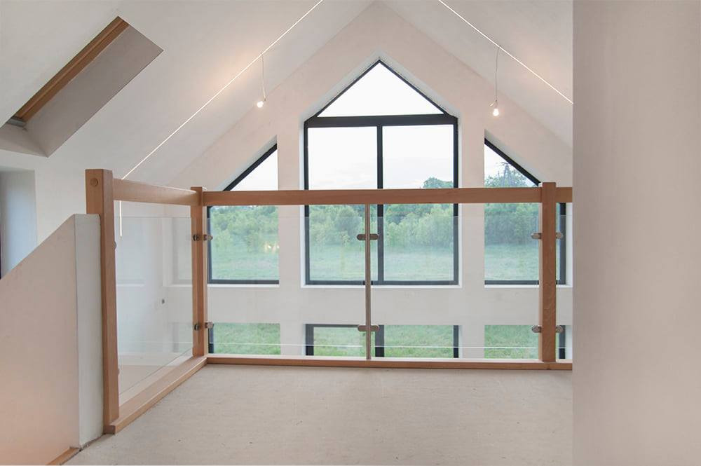 Nowoczesne wnętrze poddasza z drewnianą balustradą z elementami szklanymi, widok na zieleń przez okna o nieregularnym kształcie.