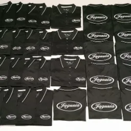 Partia czarnych koszulek polo z białym logo 'Jagusia' ułożona na białym tle, widoczne detale wykończenia kołnierzyka i plisy guzikowej.