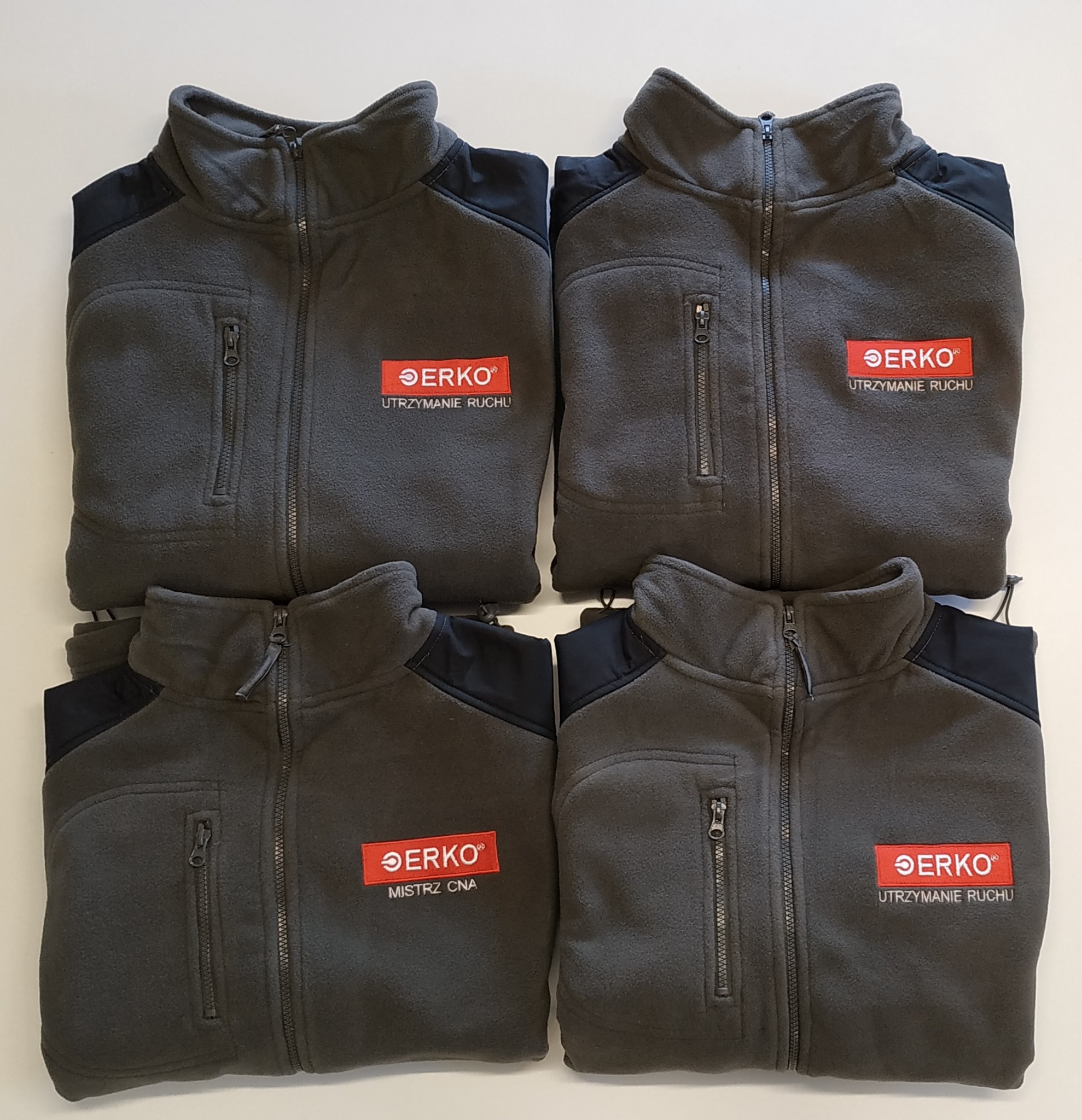 Cztery szare polarowe bluzy robocze z czarnymi wstawkami na ramionach i wyhaftowanym logo firmy ERKO z różnymi napisami: Utrzymanie Ruchu oraz Mistrz CNA.