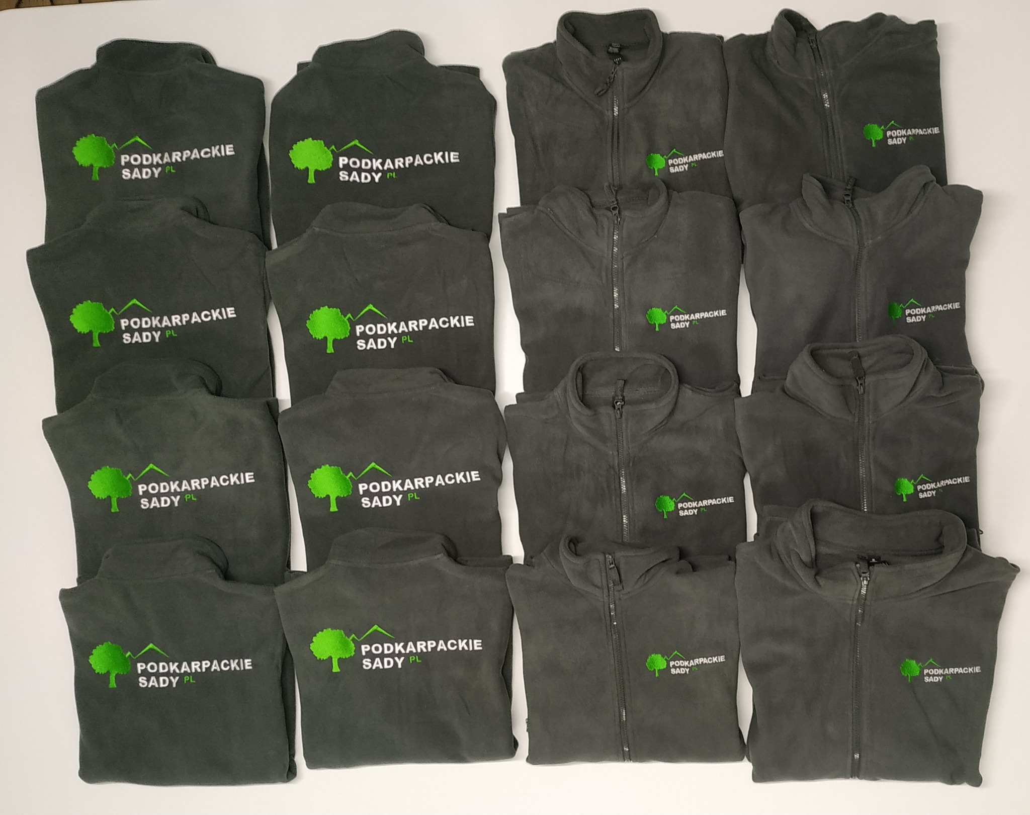 Stos polarowych bluz z wyhaftowanym logo 'Podkarpackie Sady PL' z motywem zielonego drzewa i gór, ułożonych na białym tle.