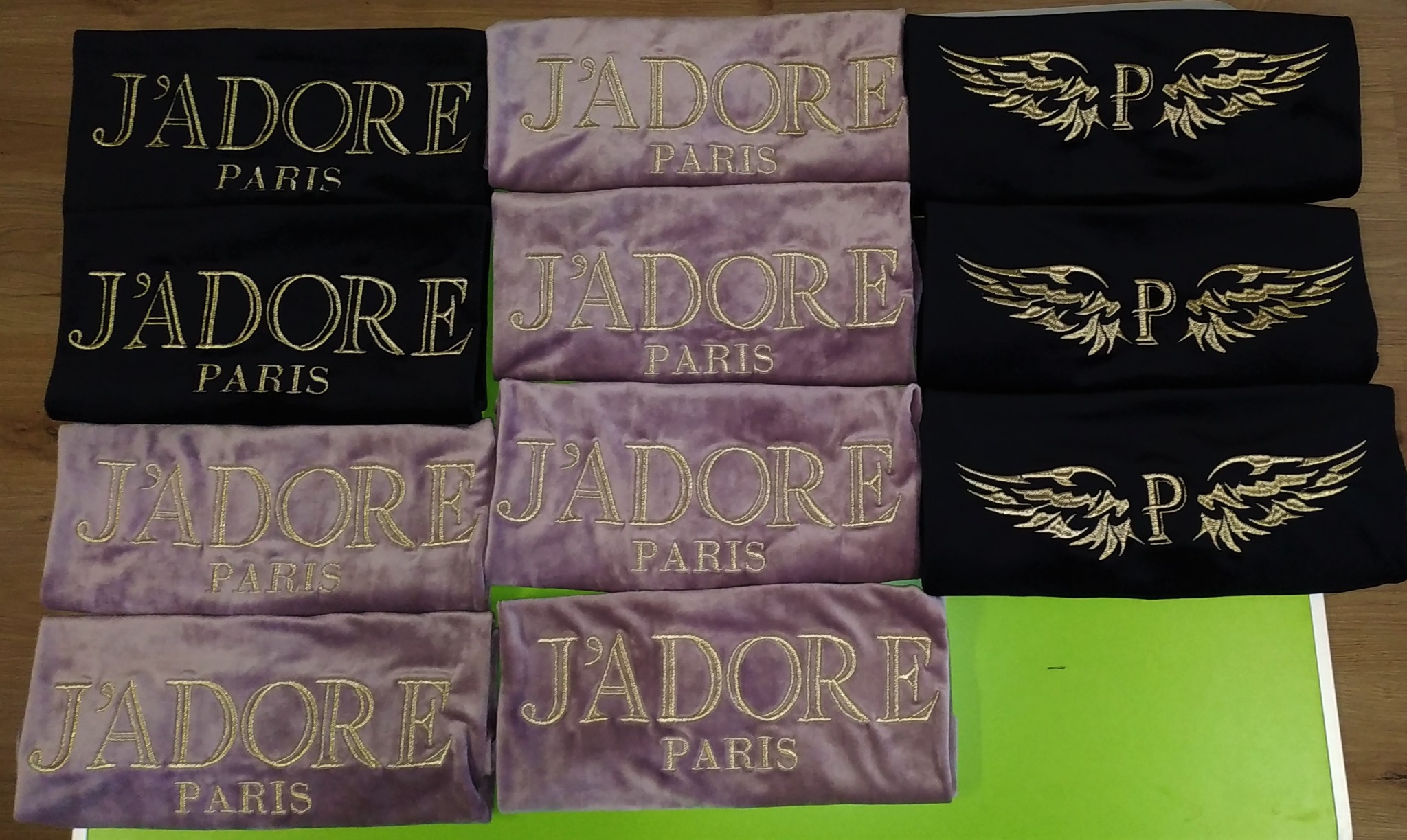 Stos ubrań z haftowanymi napisami 'J'ADORE PARIS' w kolorze złotym na czarnym i liliowym tle oraz czarne ubrania z haftowanym motywem skrzydeł i litery 'P'.
