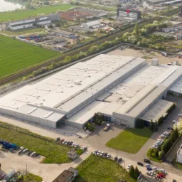 Szeroki kadr z lotu ptaka przedstawiający kompleks hal przemysłowych z blachy trapezowej, otoczonych zielenią pól i zabudową miejską.